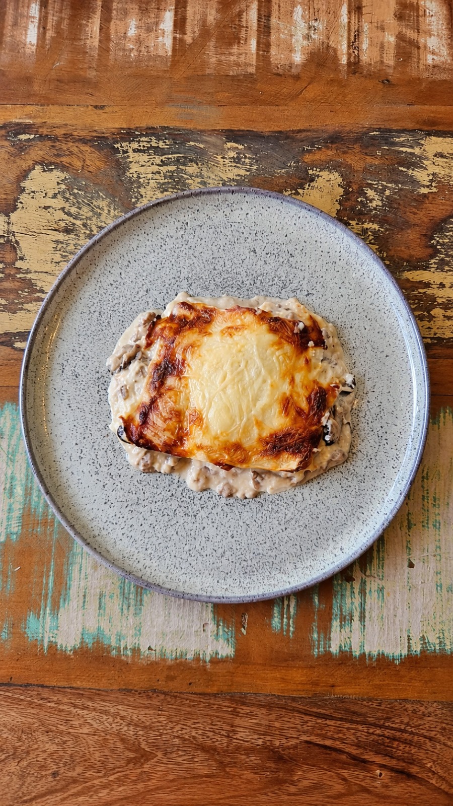 Lasanha de funghi com gruyère servida em prato cinza sobre mesa rústica.